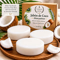 a set of white coconut soap bars with a box of the same soap bars, along with some coconut pieces and a palm leaf.