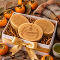 A white box containing three round, brown soap bars with a gold ribbon bow, surrounded by oranges and a cozy blanket on a wooden surface.